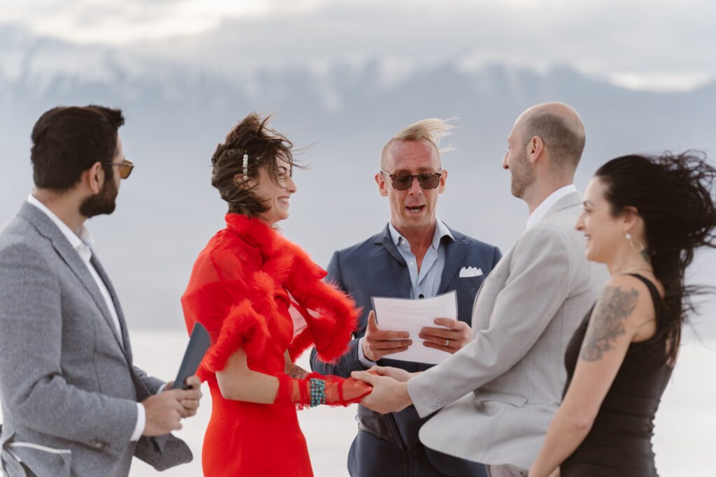 A couple says vows surrounded by friends on a salt flat on a very windy day. They and their guests are laughing