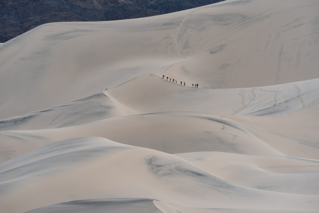 People hike up huge sand dunes. The people look very small.