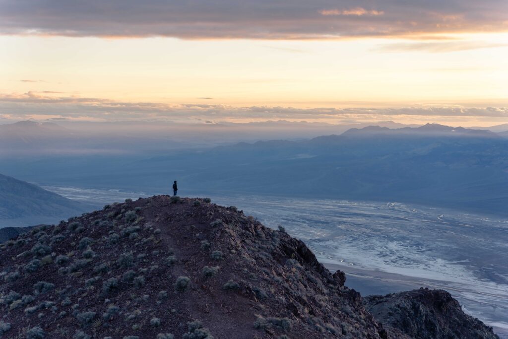 A person stands on a mountain looking out over a valley of salt as the sun sets