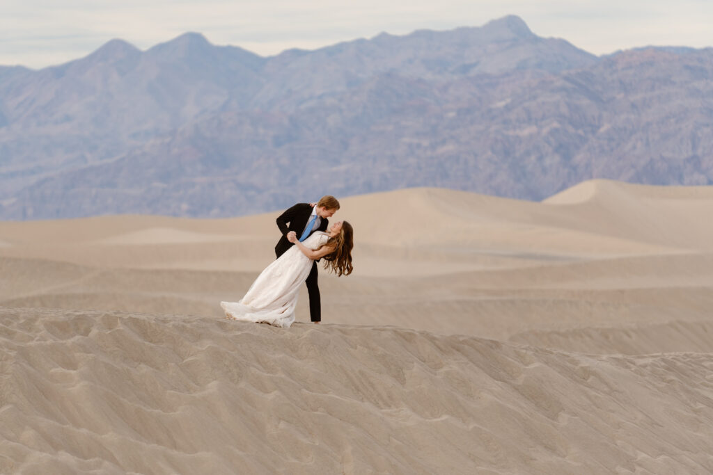 A man in a suit dips a woman in a wedding dress on top of a sand dune in Death Valley