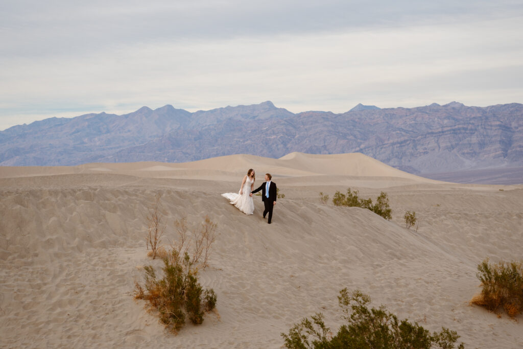 A couple in wedding clothes walk over sand dunes on a cloudy day in Death Valley