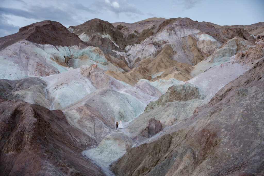 A couple in wedding clothes holds hands and walks through colorful hills of Artist Palette