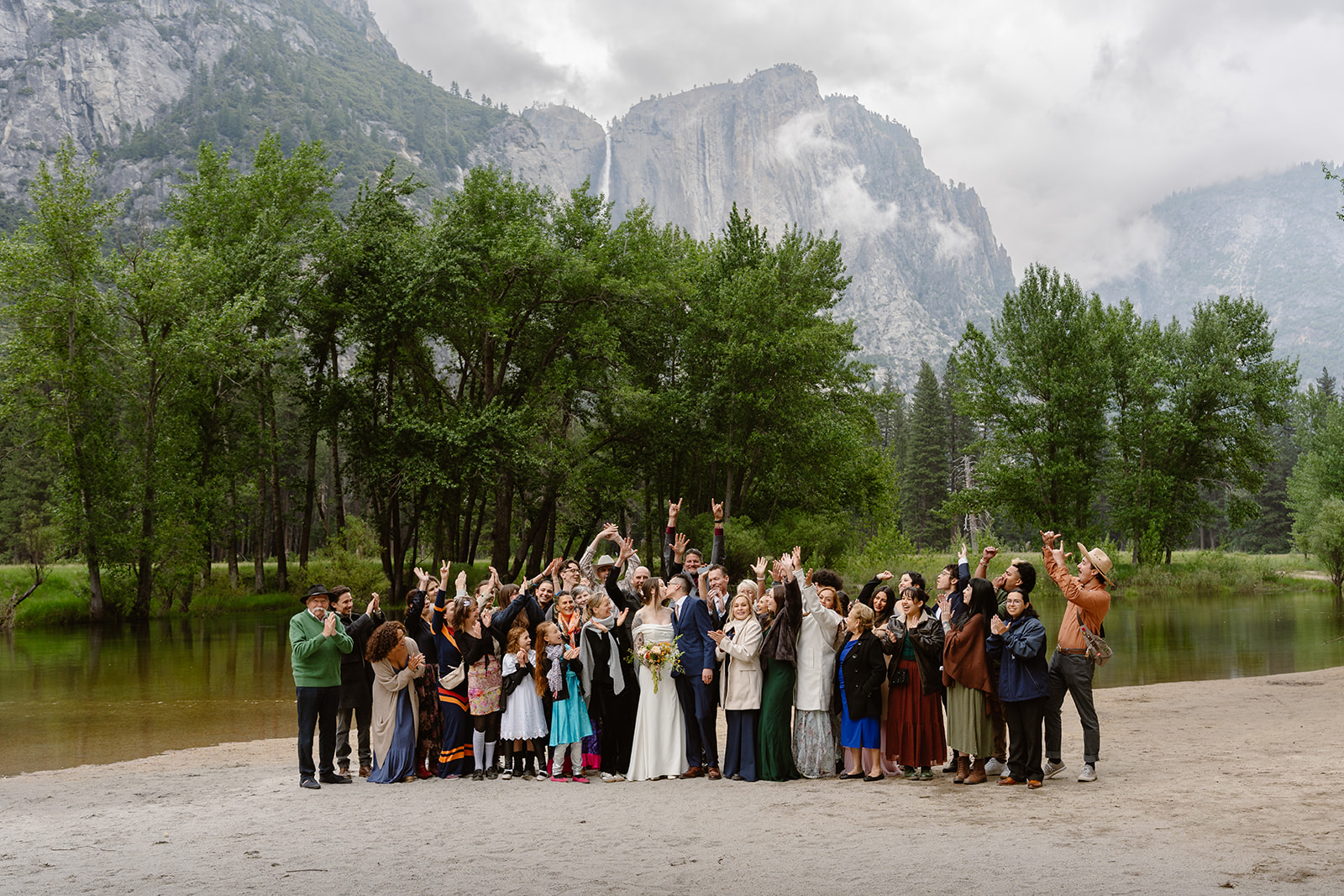 A newlywed couple and their guests cheer at the end of their elopement ceremony on a riverside beach in Yosemite