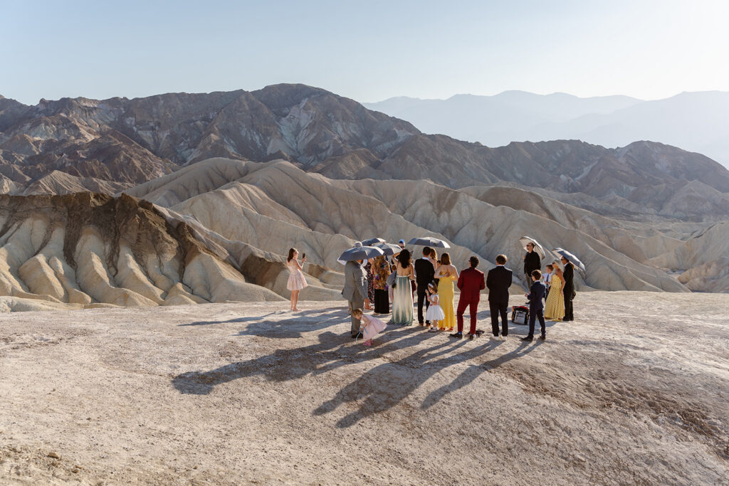 a group of guests huddle under sun umbrellas in the desert during an elopement ceremony
