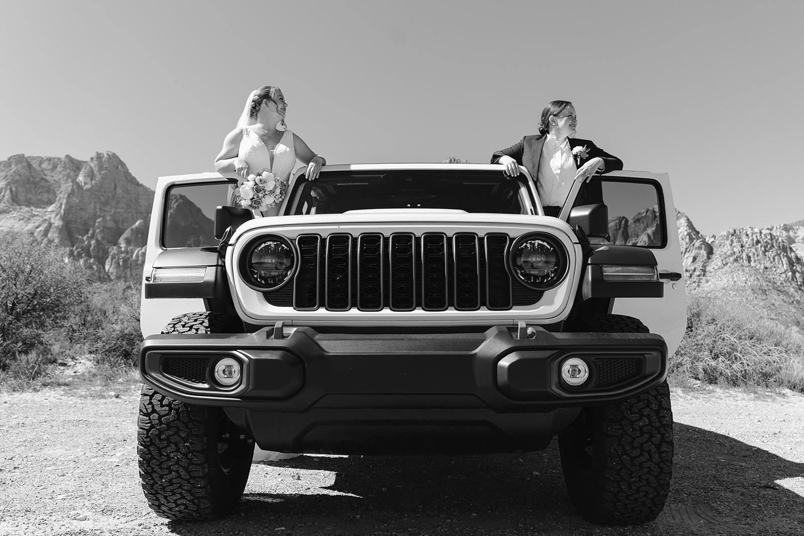 A Jeep with the front doors open, two women standing up inside the car looking out into the distance in their wedding clothes