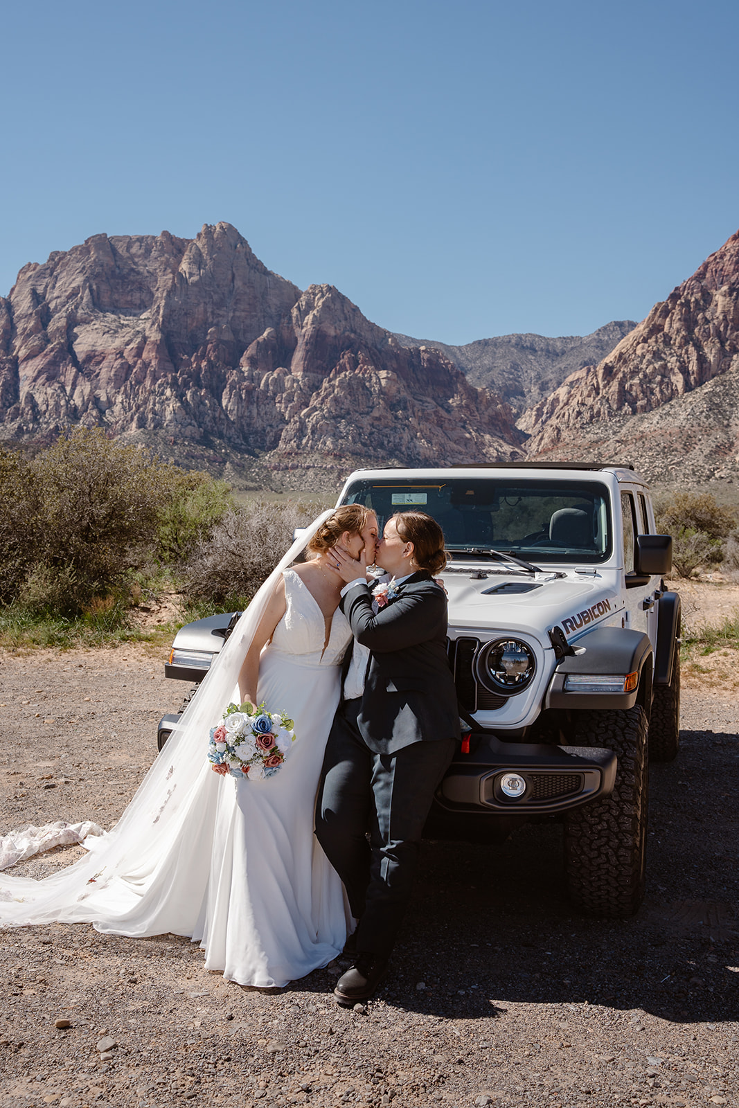 Two women kiss while seated on the front bumper of a white Rubicon Jeep. Red Rock Canyon is in the background