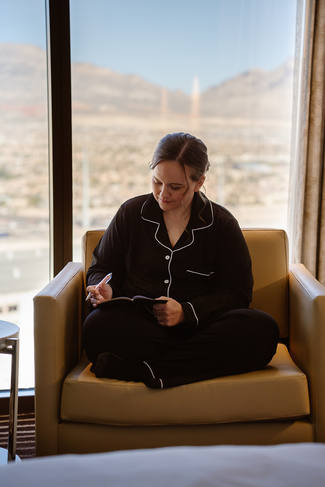 a woman sits in her hotel room writing private vows in her pajamas. the desert is visible behind her through the window