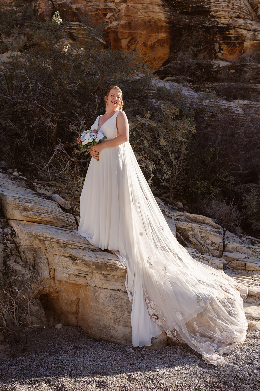 A bride stands on a rock smiling as her wedding dress cascades over the side and the sunlight shines through it creating a lovely glow
