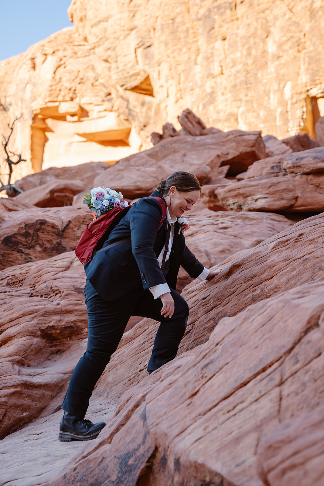 A bride in a suit and a red backpack with a bouquet sticking out climbs a large rock