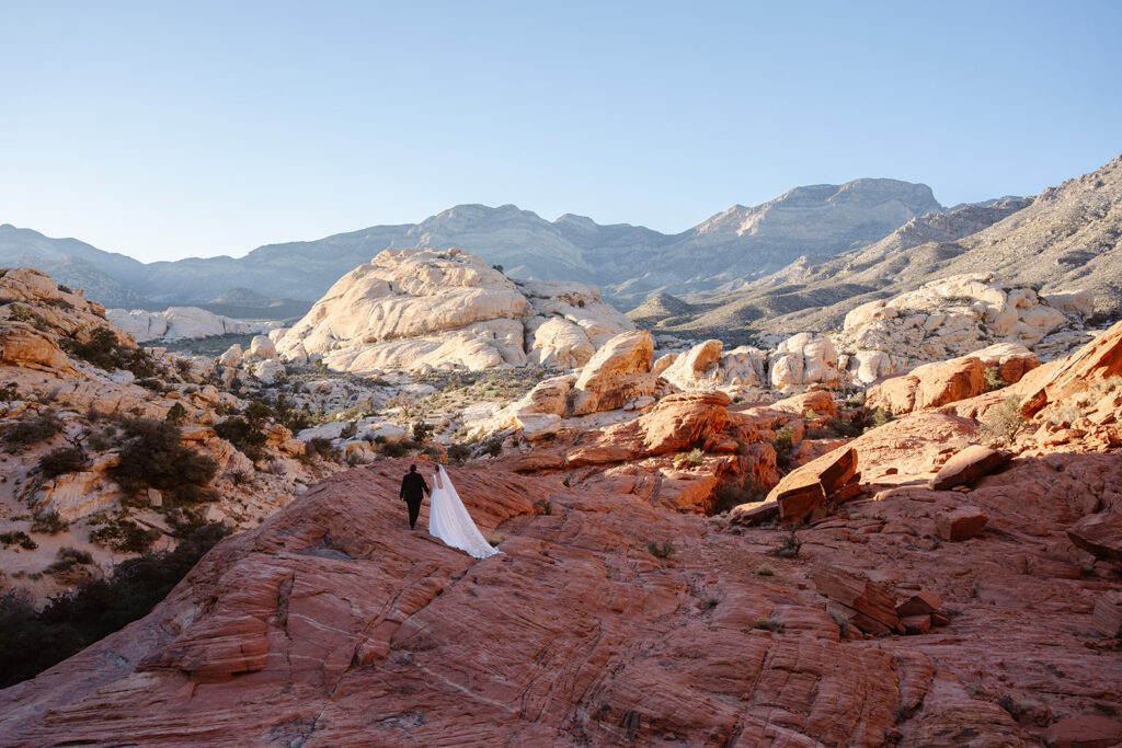 Two brides walk hand in hand over red rocks as the sun sets over the desert landscape behind them in red rock canyon