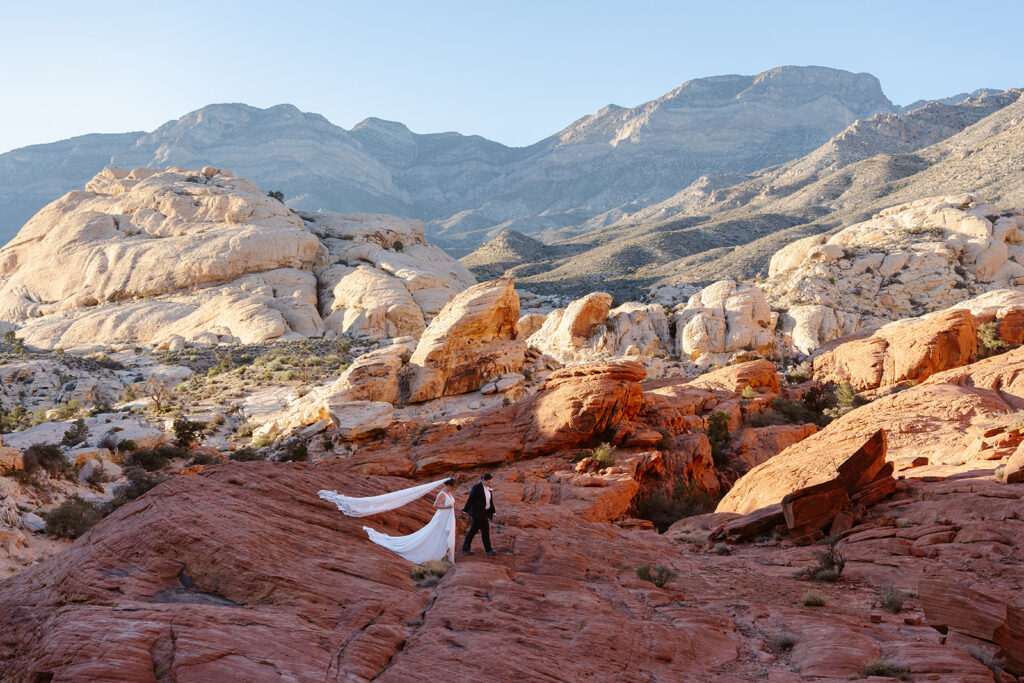 Two women dressed in wedding clothes walk over red rocks after their queer elopement in Red Rock Canyon