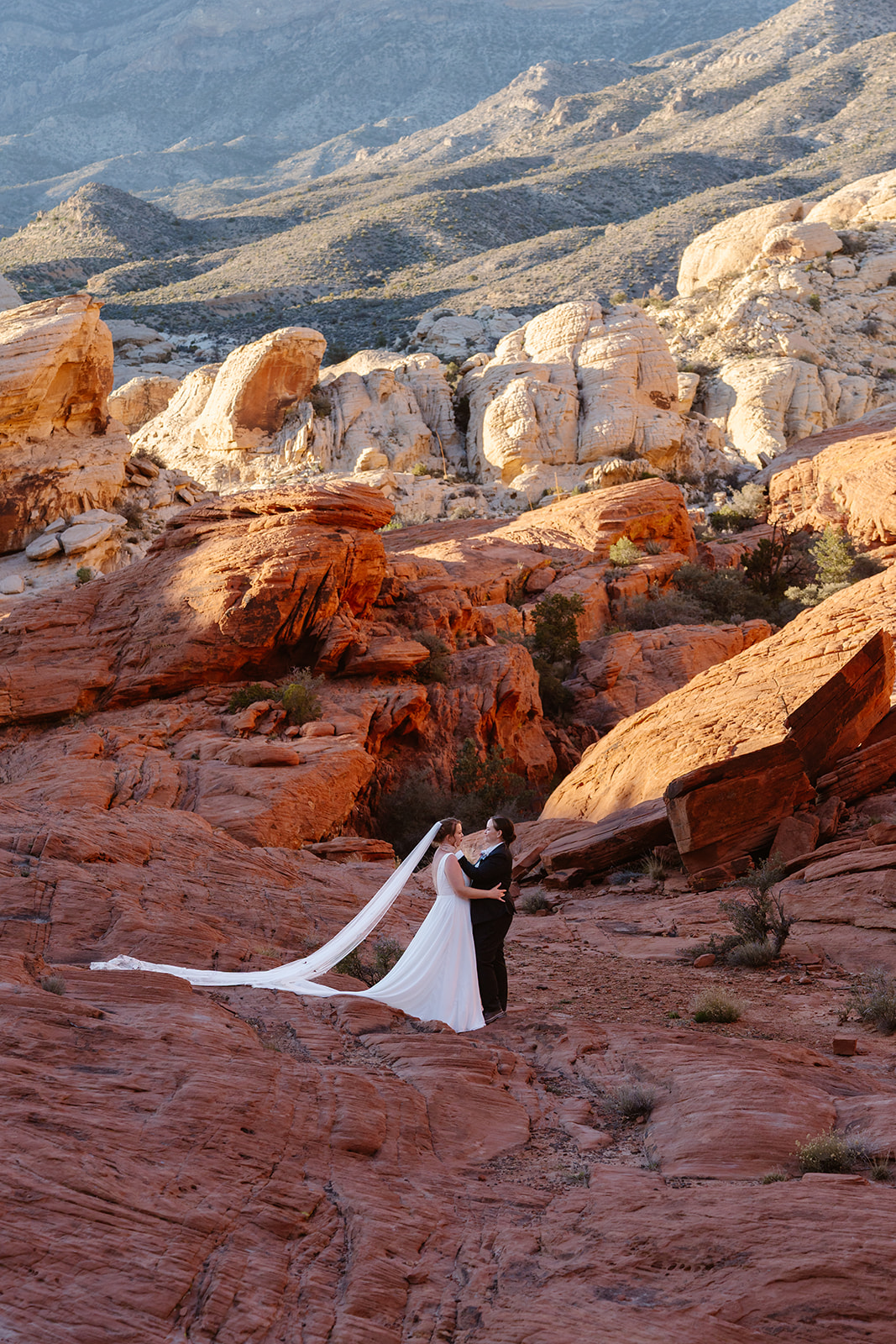 two women embrace amongst red rocks as the sun sets on the desert landscape behind them