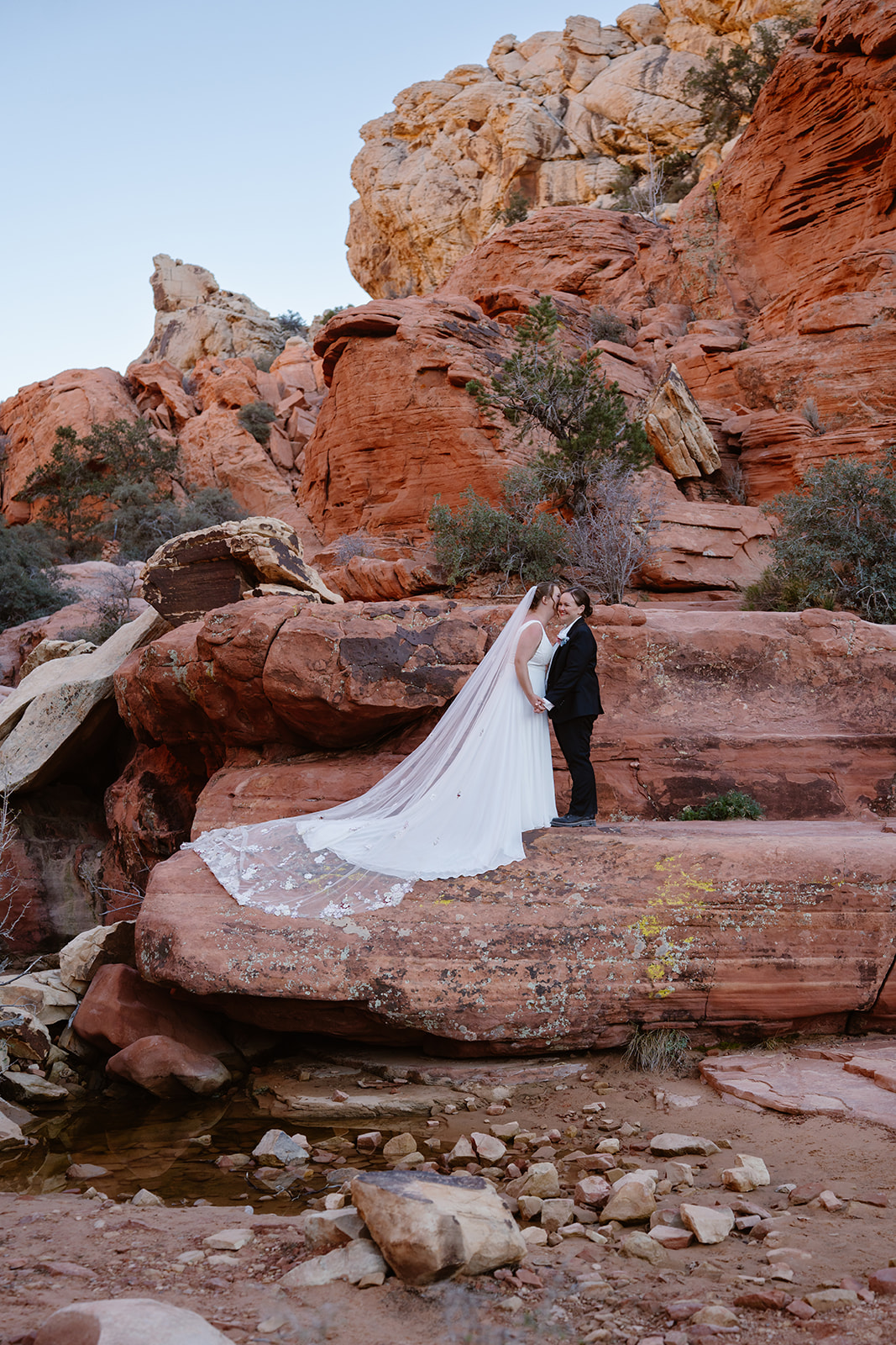 One bride laughs as her new wife whispers something in her ear. They stand amongst red rocks and a creek