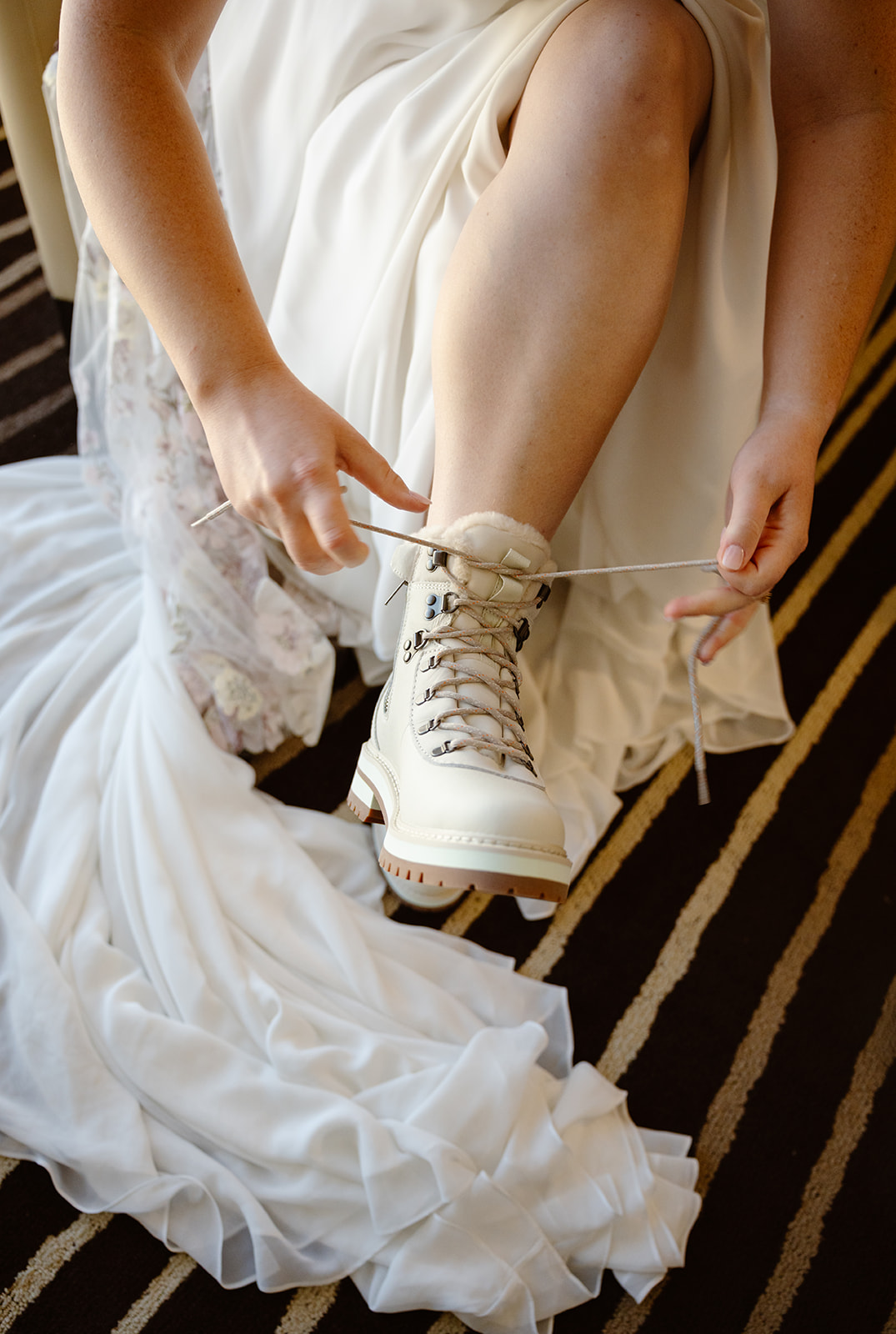 a bride laces up white hiking boots among the folds of a long white dress