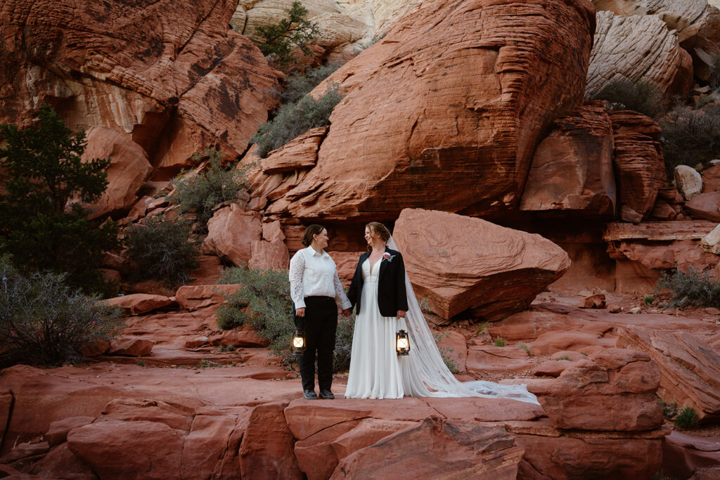Two brides hold hands and look at each other smiling while holding lantern as the sun goes down after their queer elopement in red rock canyon