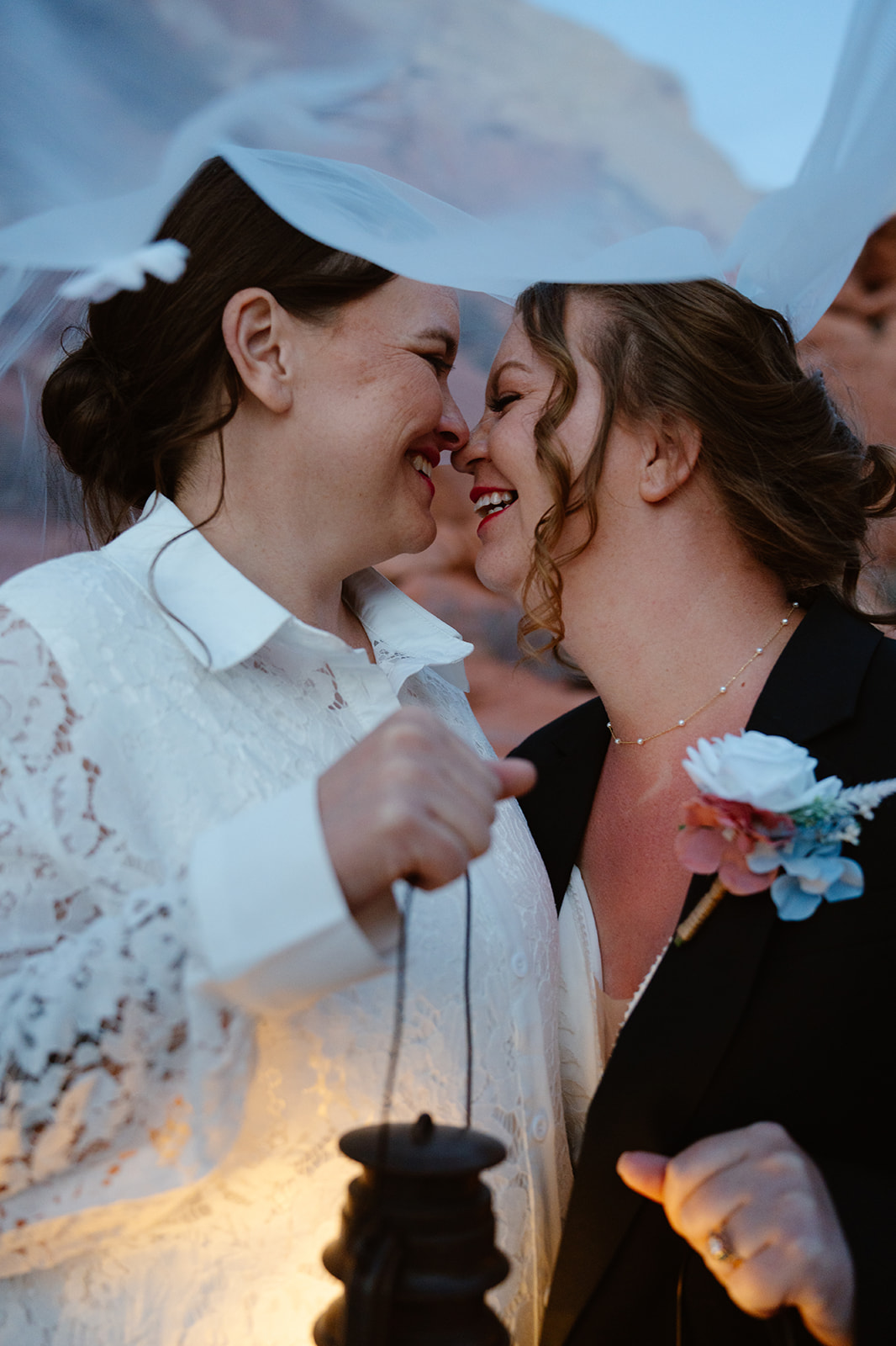 Two brides laugh and nuzzle each other underneath a veil, holding lanterns
