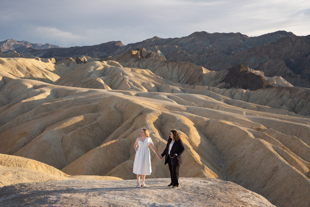 Two brides hold hands looking into the distance among tan colored hills in Death Valley