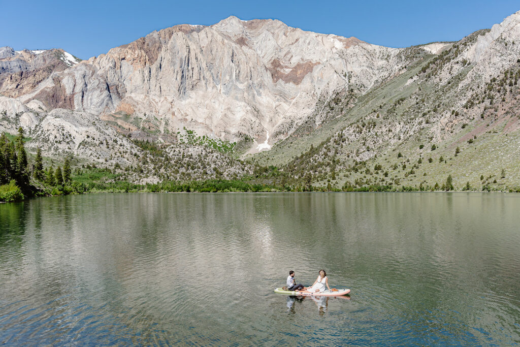 A couple sits on a paddleboard floating in the middle of a lake with a large mountain rising up behind them in the background