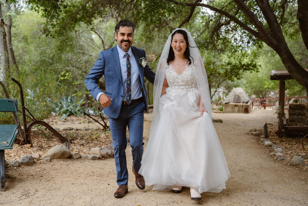 A bride and groom walk toward the camera smiling. There are trees and a dirt path in the background