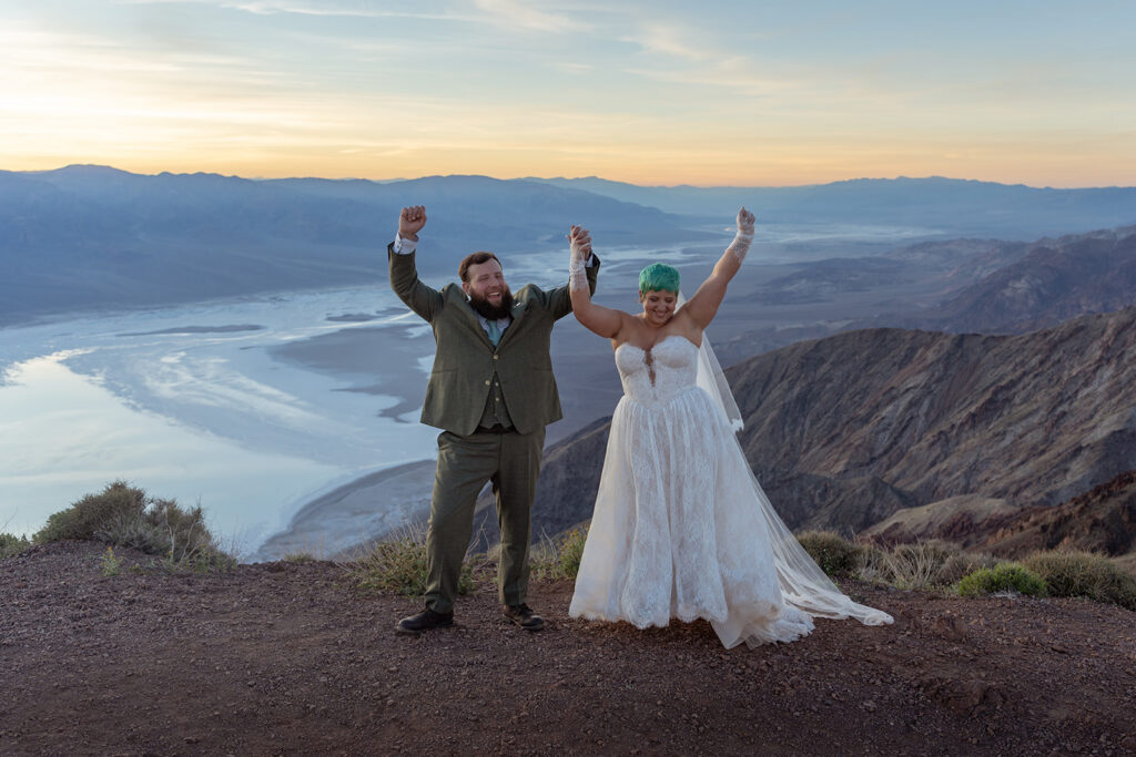 A bride and groom jump in the air smiling and holding hands. A valley full of salt stretches out behind them